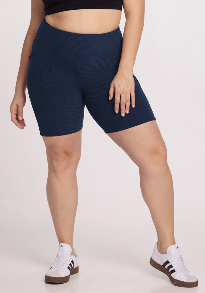 Wearing Woolx Miley Bike Shorts in Deep Navy, a black top, and white sneakers stands against a light background.