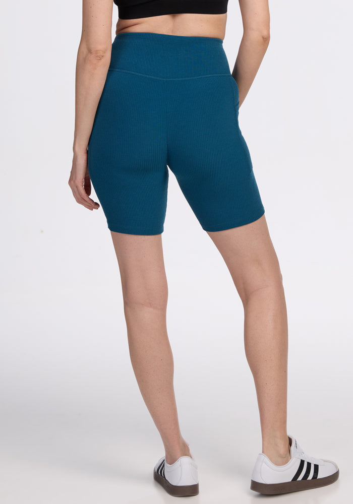 Woman wears Woolx Miley Bike Shorts in Moroccan Blue, a black crop top, and white sneakers, standing with back to camera.