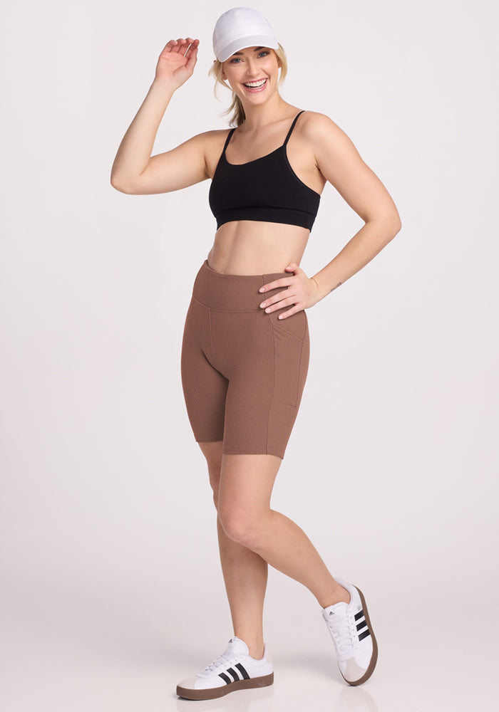 Woman in Miley Bike Shorts by Woolx, black sports bra, white cap, and sneakers smiles against a plain background.