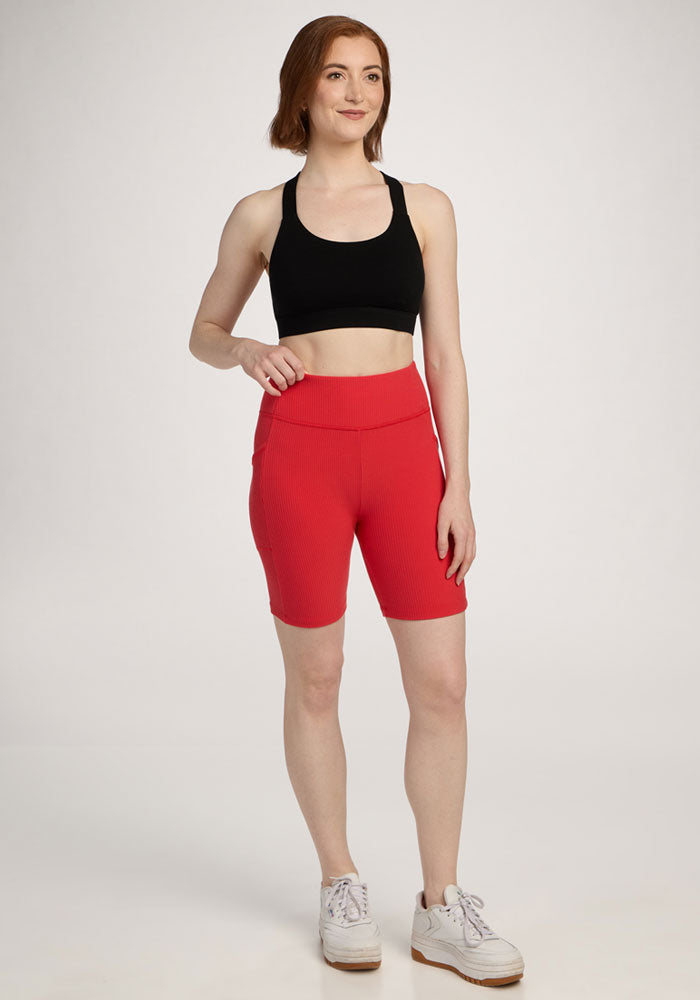 Woman with short red hair smiles in a black sports bra and Woolx Miley Bike Shorts - Hibiscus, plus white sneakers.