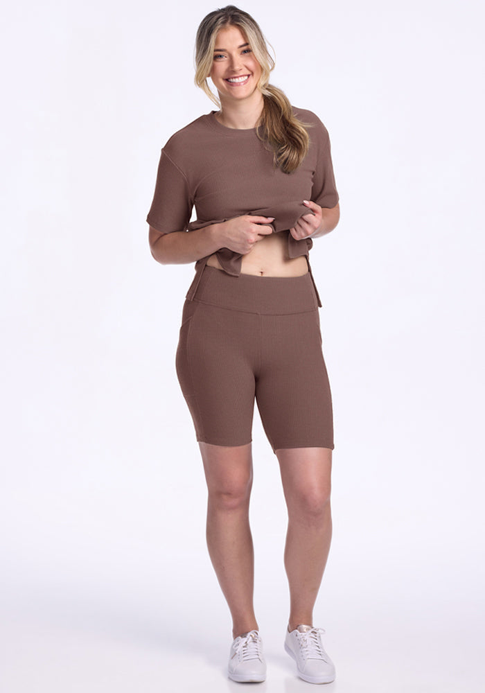 Smiling woman in Woolx Miley Bike Shorts - Peppercorn, lifting her top slightly against a white background.