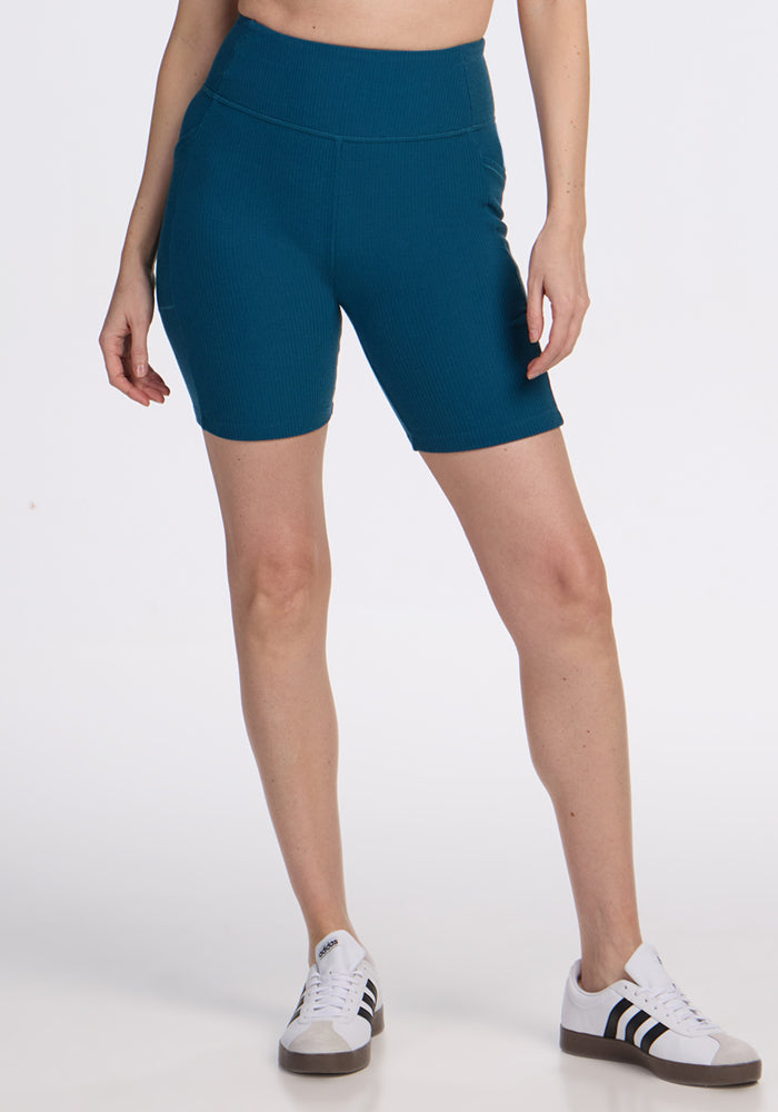Person wears Woolx Miley Bike Shorts in Moroccan Blue with white sneakers, standing against a plain white background.