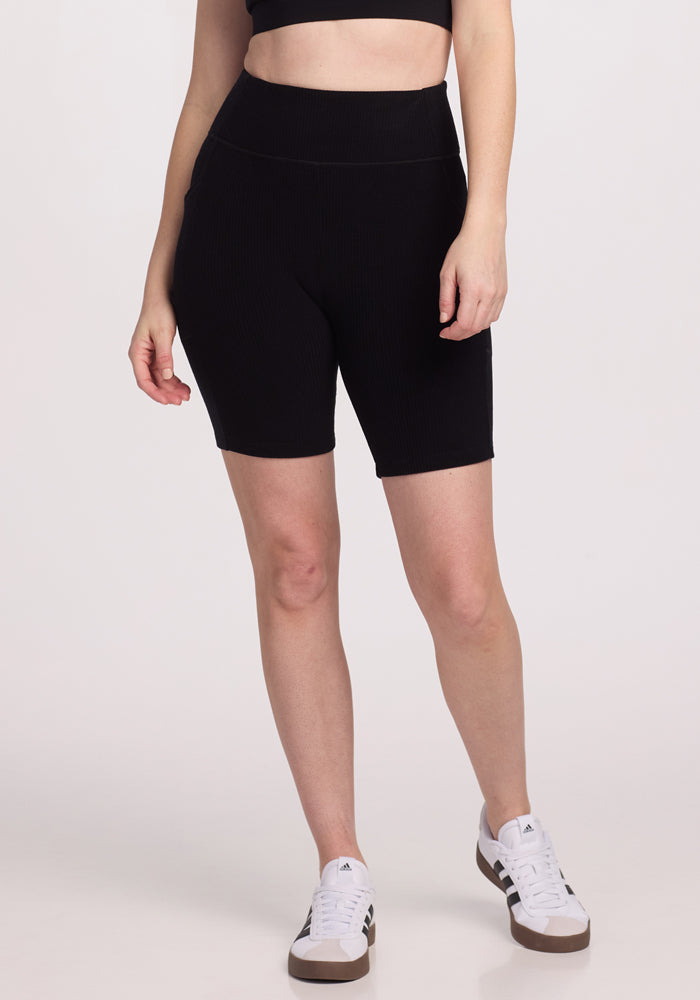 A person is wearing the Woolx Miley Bike Shorts in black along with a matching black crop top. They stand with one hand by their side, also sporting white sneakers with black stripes against a plain white background. | Lilly is 5’9”, wearing a size S