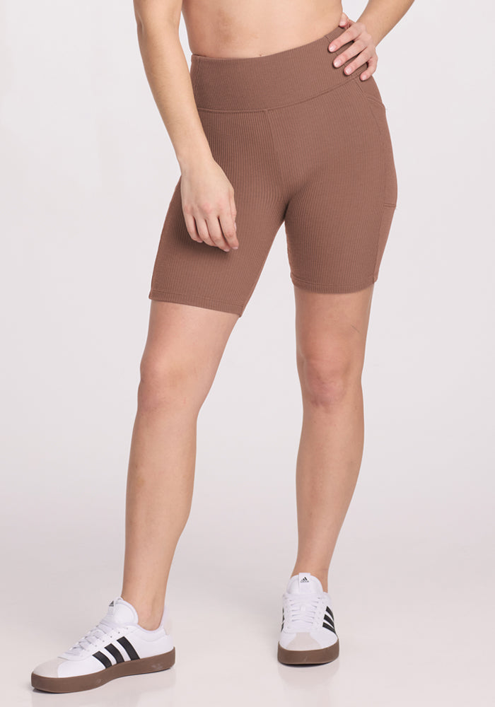 A person wears Woolx Miley Bike Shorts in Sienna Kiss and white sneakers with black stripes, against a white background.