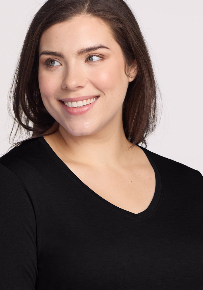 A person with long brown hair smiles while wearing the Woolx Millie Long Sleeve - Black, made from moisture-wicking material. They gaze to the left against a light, neutral background.