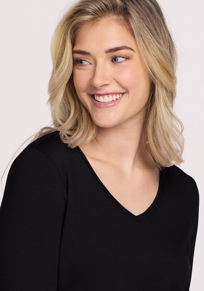 A person with shoulder-length blonde hair and light skin smiles, gazing sideways. They are wearing the Woolx Millie Long Sleeve in black, a V-neck top featuring temperature-regulating, moisture-wicking fabric. The background is plain and light-colored.