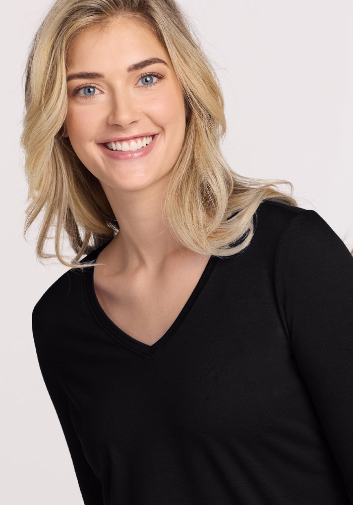A person with long blonde hair smiles while wearing a Woolx Millie Long Sleeve in black, crafted from moisture-wicking Merino wool. The background is a plain, light color.