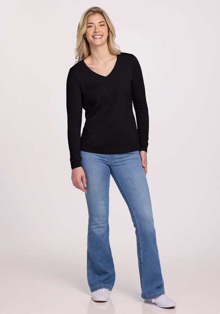 A person with long blonde hair is smiling against a plain background, dressed in Woolx's black Millie Long Sleeve top, blue jeans, and white sneakers.