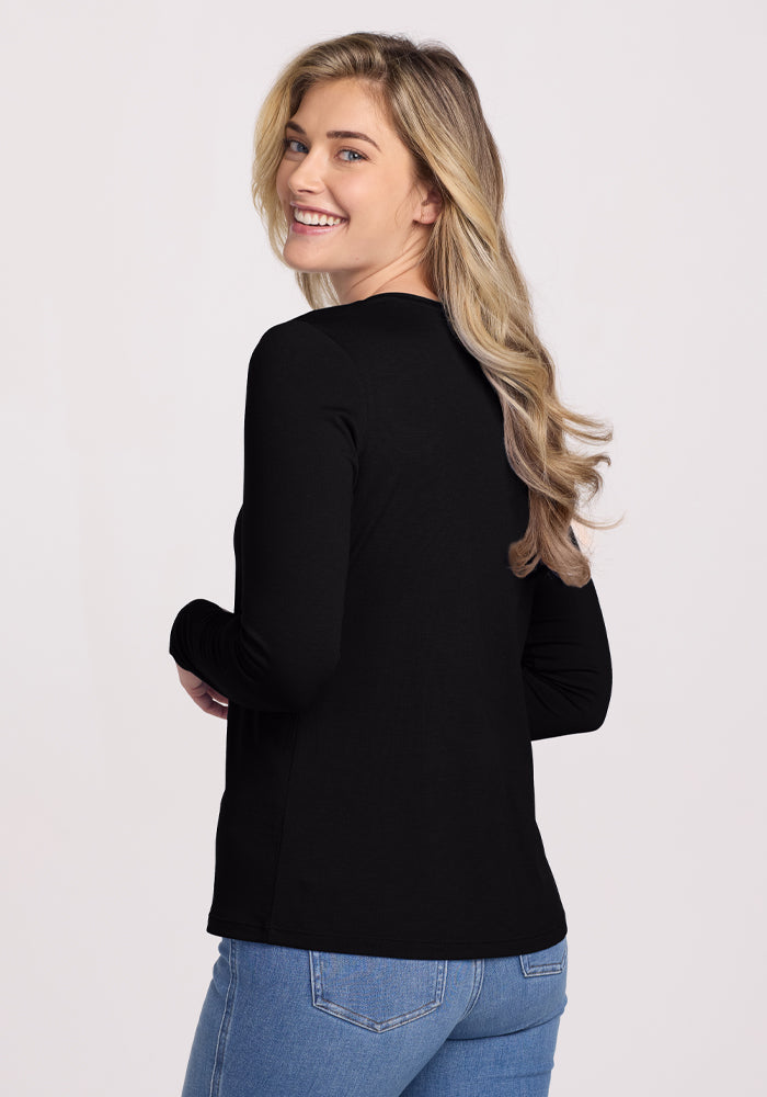 A smiling person with long blonde hair wears the Woolx Millie Long Sleeve in black and blue jeans, standing slightly turned towards the camera against a plain background.