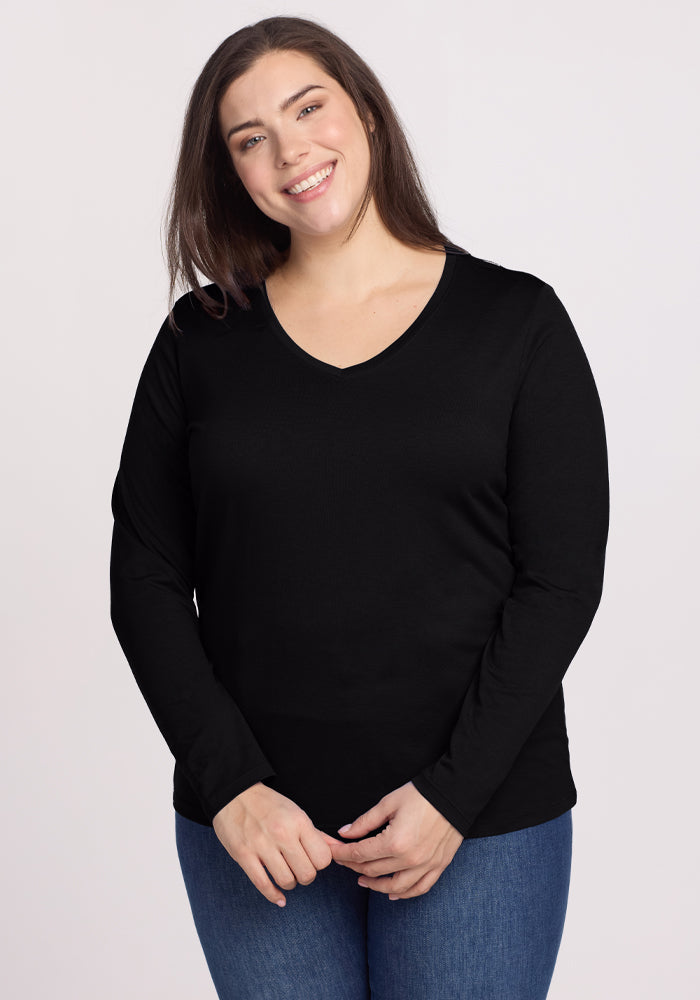 A person with long brown hair smiles wearing the Woolx Millie Long Sleeve - Black and blue jeans, standing against a plain light background, effortlessly combining comfort and style. | Juliana is 5'10", wearing a size XL