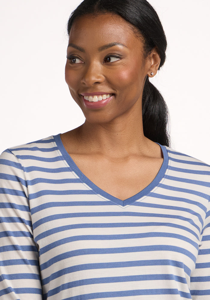 A woman with long dark hair in a low ponytail smiles and looks to the side, wearing the Woolx Millie Long Sleeve in Blue Sand Stripe. The background is plain and light-colored.