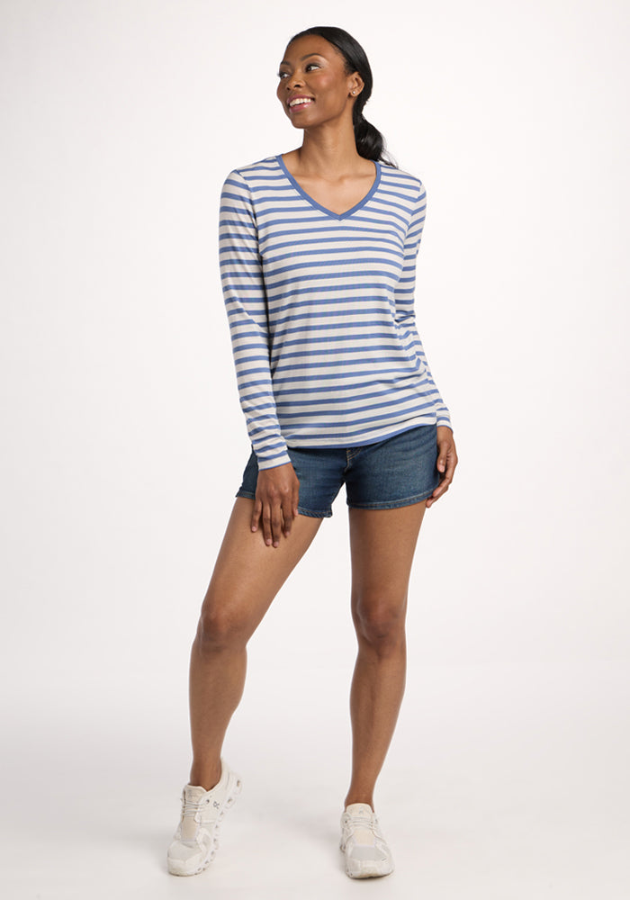 A woman smiles, wearing the Woolx Millie Long Sleeve in Blue Sand Stripe, paired with denim shorts and white sneakers, set against a plain light background.