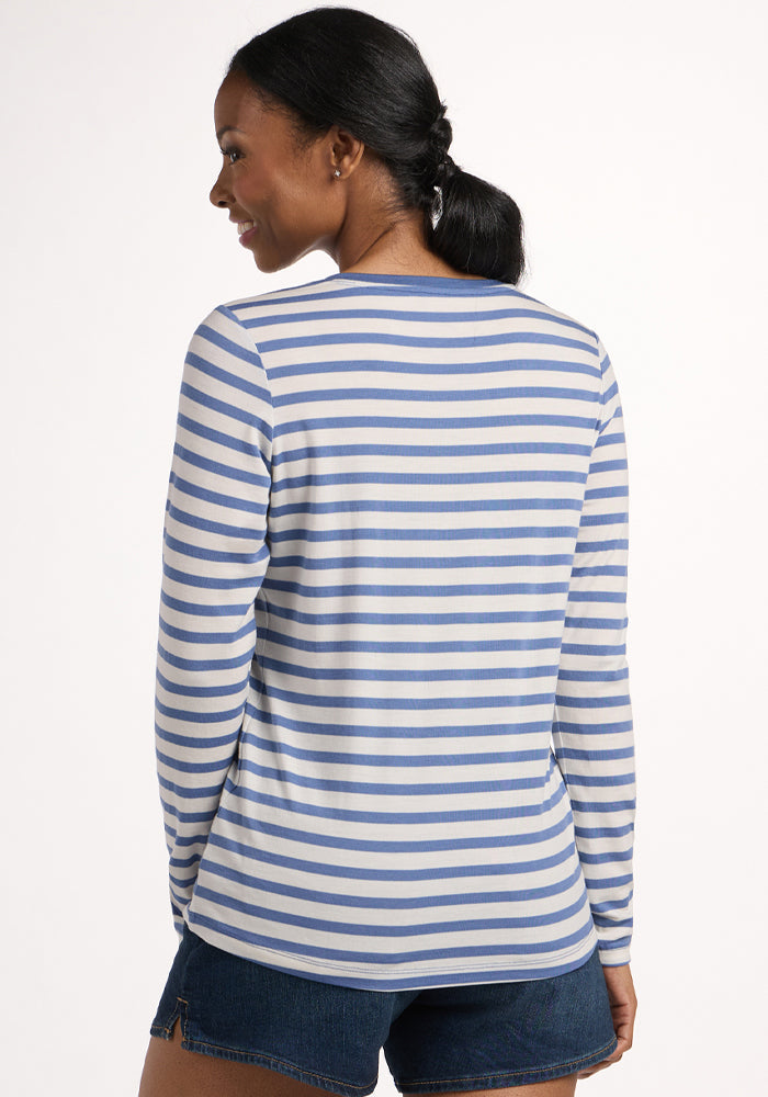 A woman with a low ponytail faces away, wearing the Woolx Millie Long Sleeve in Blue Sand Stripe and denim shorts. This moisture-wicking, temperature-regulating Merino wool top embodies effortless style against a plain light background.