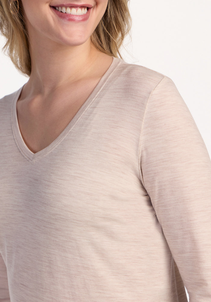 A person smiles from the shoulders up, wearing the Woolx Millie Long Sleeve in Cream Heather, a travel-ready Merino wool V-neck top, set against a plain white background.
