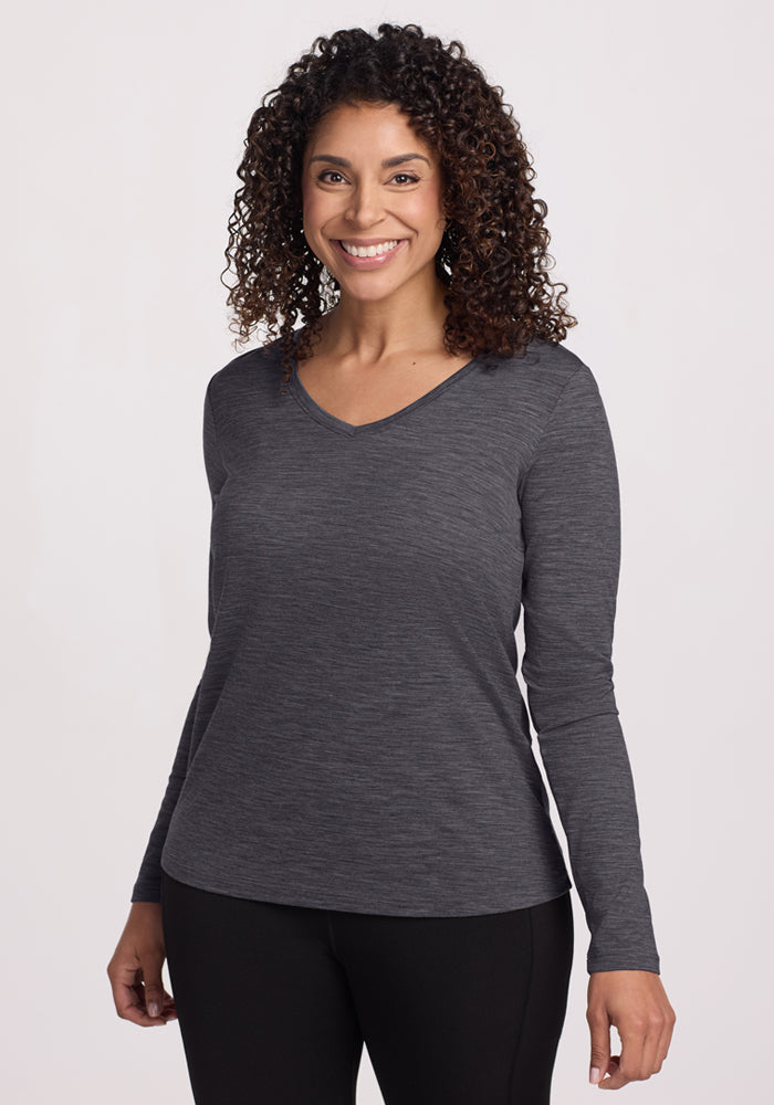 Smiling woman in Woolx Millie Long Sleeve, Cinder Melange, with curly hair and black pants against a light background.
