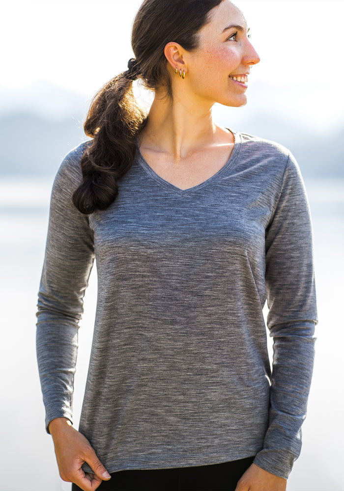 A woman with dark hair in a ponytail stands outdoors, smiling and looking to her right, wearing the Woolx Millie Long Sleeve top in Graphite Heather. The background is bright and slightly blurred.