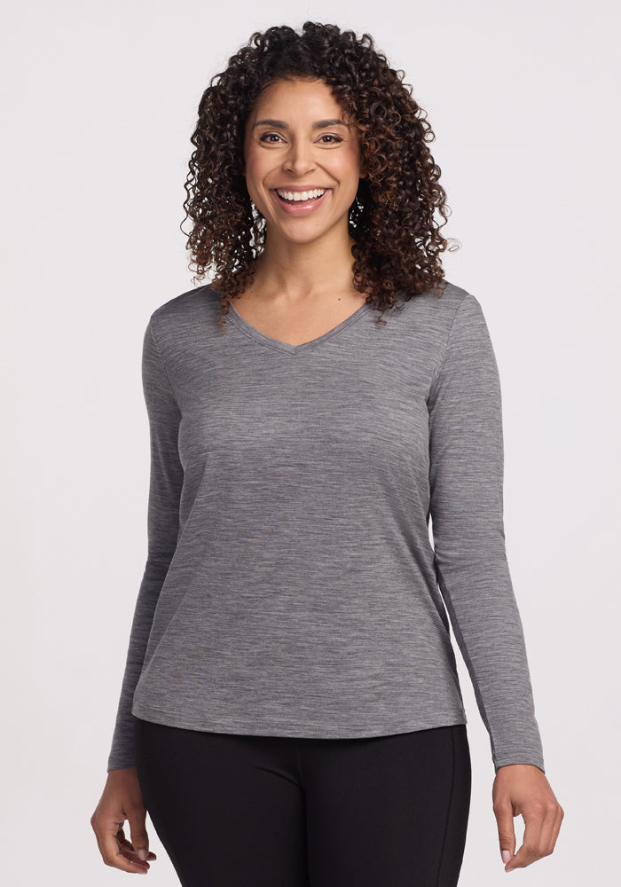 A person with curly hair is smiling in a Woolx Millie Long Sleeve in Graphite Heather and black pants, standing against a plain white background. | Kisha is 5’8”, wearing a size M