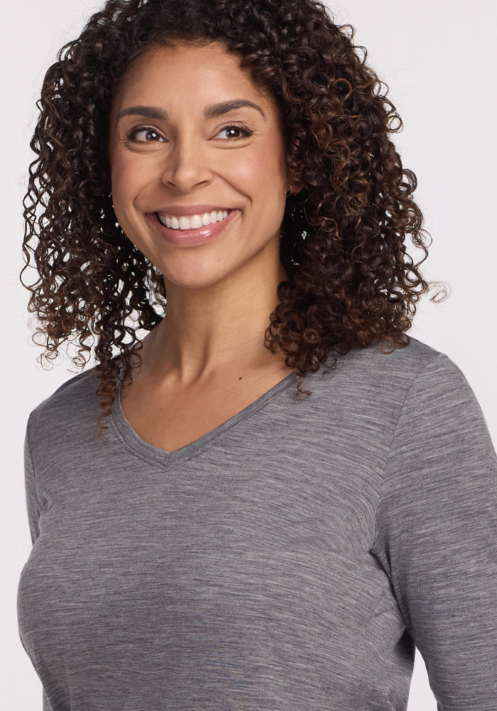 A smiling person with curly hair is seen wearing the Woolx Millie Long Sleeve in Graphite Heather, crafted from temperature-regulating Merino wool, while glancing slightly to the side against a plain background.