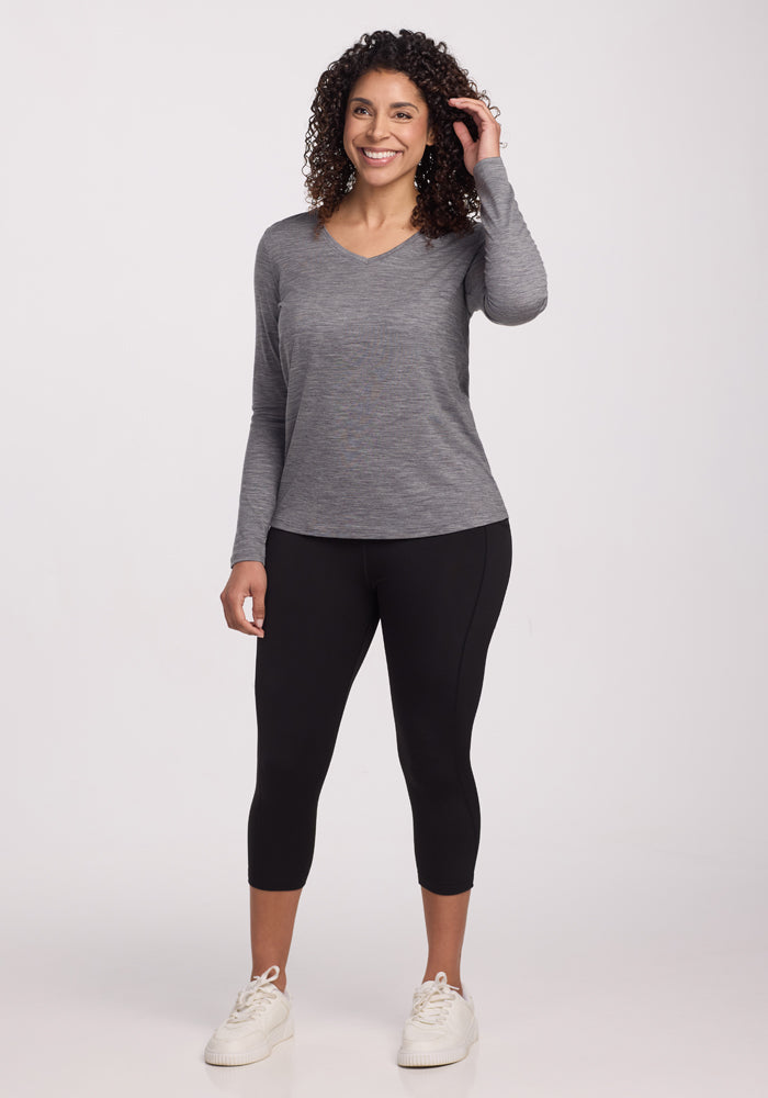 A woman with curly hair wears the Woolx Millie Long Sleeve in Graphite Heather, paired with black leggings and white sneakers, standing and smiling against a plain white background.