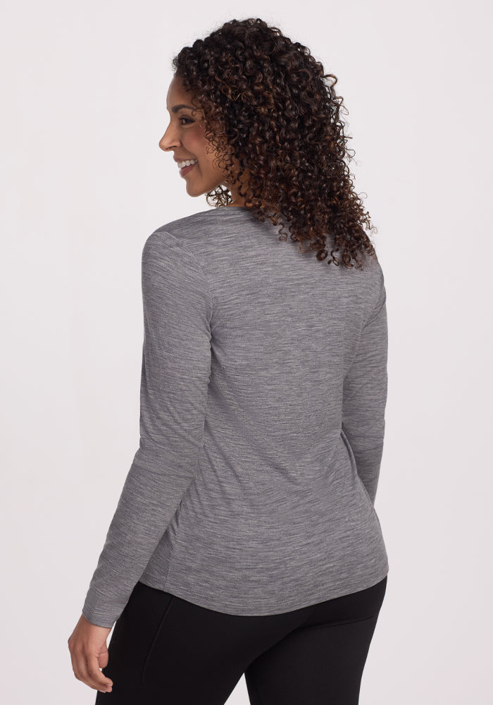 A person with curly hair wears the Woolx Millie Long Sleeve in Graphite Heather and black pants, turned slightly right and smiling while looking off-camera against a plain white background.
