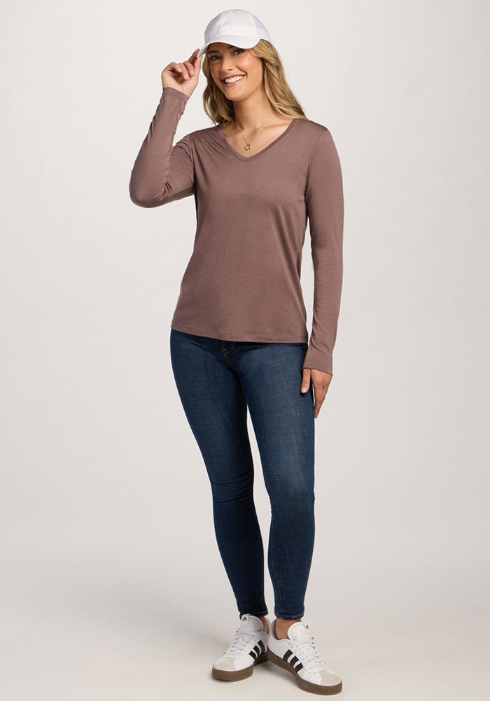 Woman smiles in a Woolx Millie Long Sleeve - Peppercorn, blue jeans, and white sneakers against a plain background.
