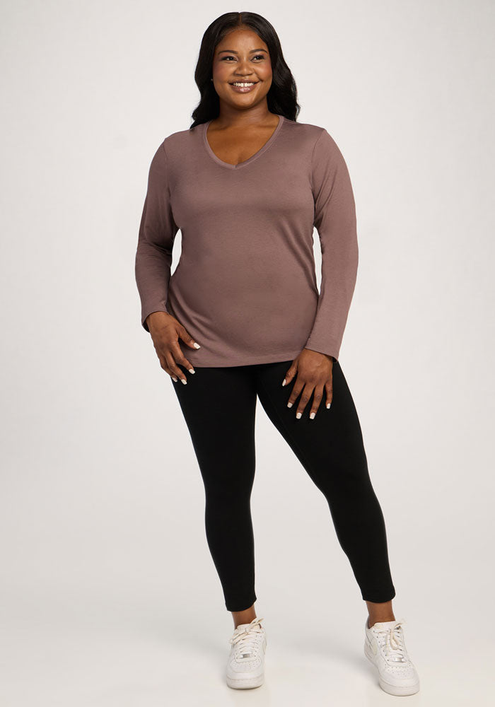 Smiling woman wears Woolx Millie Long Sleeve in Peppercorn, black leggings, and white sneakers against a plain background.