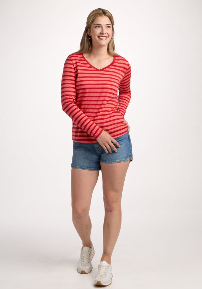 A woman smiles, wearing the Woolx Millie Long Sleeve in Wildberry Stripe—featuring moisture-wicking Merino wool—paired with blue denim shorts and white sneakers, standing against a plain white background.