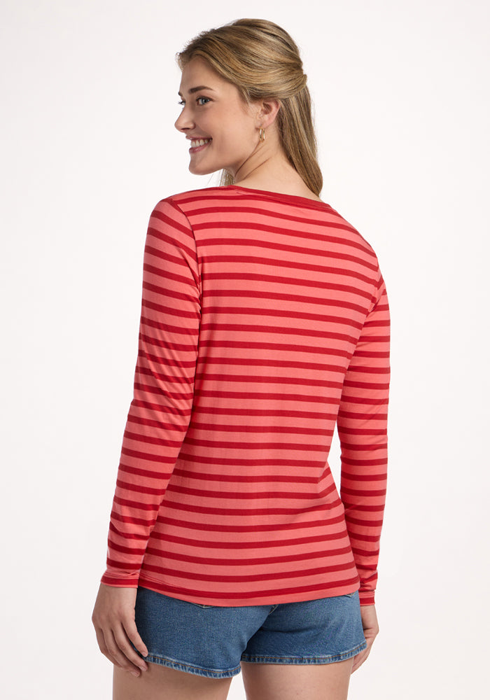 A woman with long blonde hair smiles over her shoulder, wearing the Woolx Millie Long Sleeve - Wildberry Stripe, a temperature-regulating, moisture-wicking Merino wool top with red and pink stripes, plus denim shorts, against a white background.