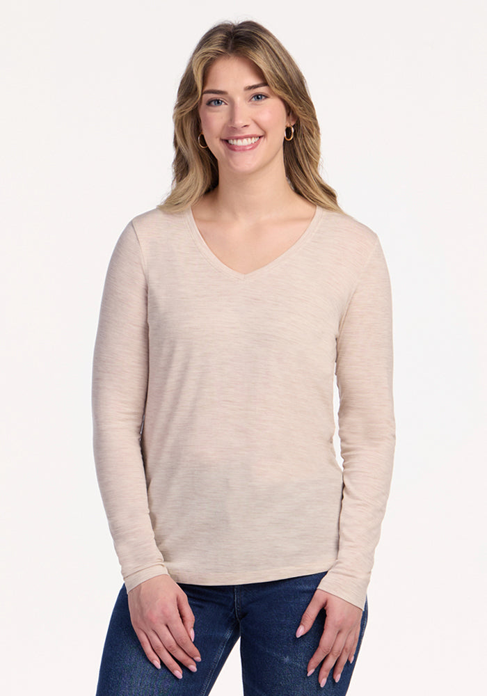 A woman with light brown hair smiles against a plain white background, wearing the Woolx Millie Long Sleeve in Cream Heather and blue jeans—travel-ready and perfect for temperature regulation. | Ariana is 5'11", wearing a size S