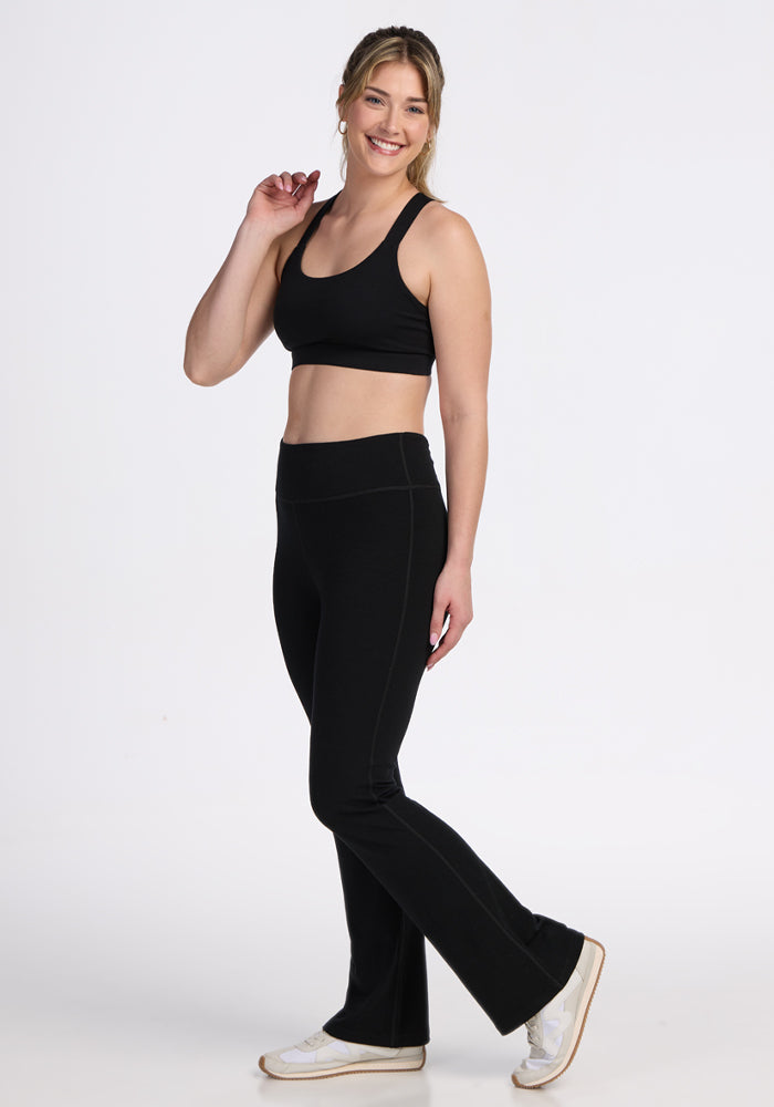 A woman stands smiling against a white background, wearing Woolx Morgan Flare Pants in black with a black sports bra and white sneakers. One hand touches her hair as she poses confidently.