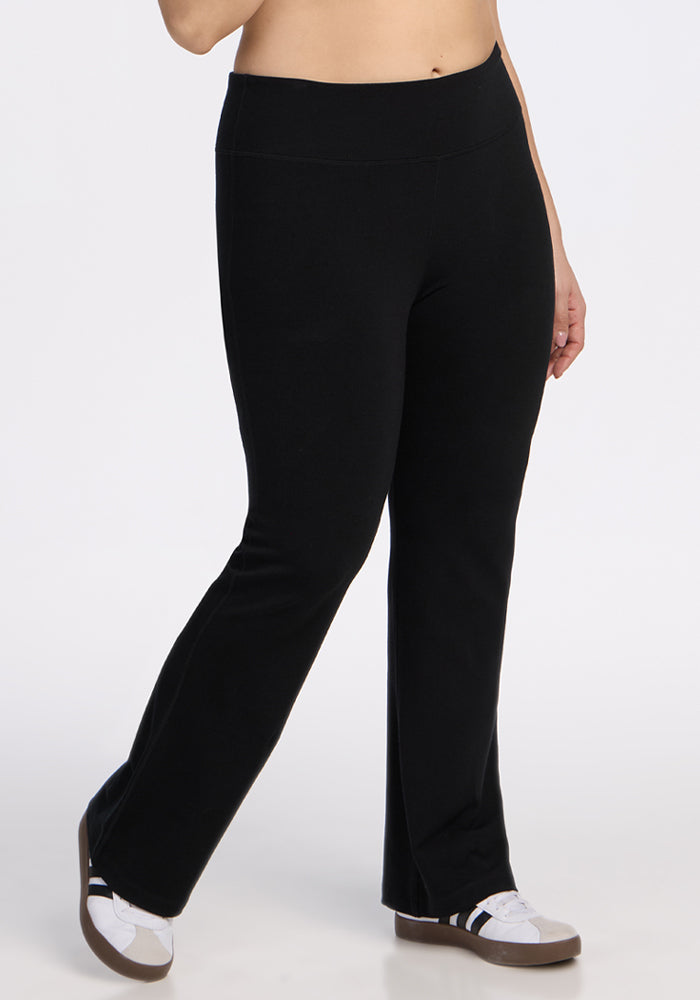 A person models Woolx Morgan Flare Pants in black against a plain white background, paired with white sneakers featuring black stripes. The image is cropped to display only the lower torso and legs.