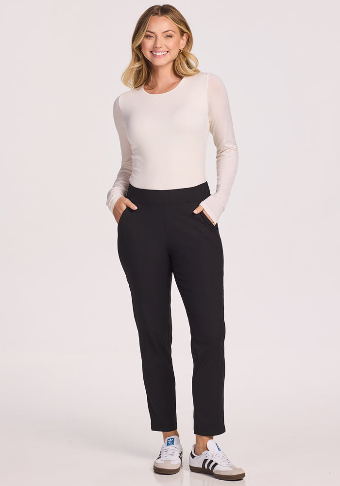 A woman smiles with her hands in her pockets, wearing Woolx Nicole Pants in black and a long-sleeve white top, paired with white sneakers featuring brown details, set against a plain light background.