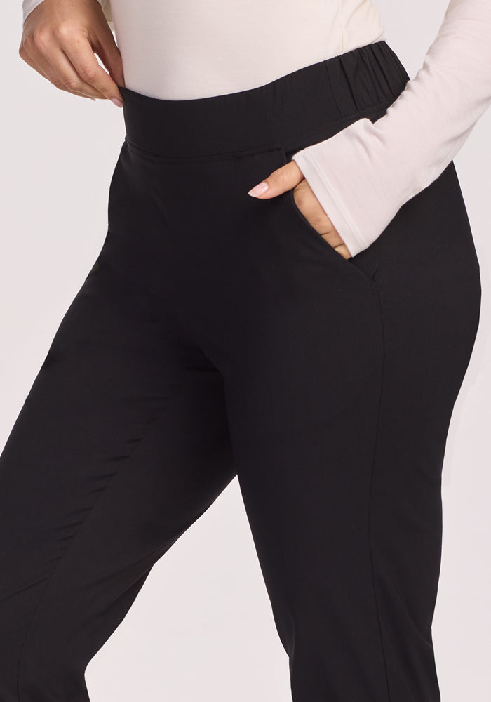 A person wears Woolx Nicole Pants in black and a long-sleeve white top with thumbholes, standing with one hand in their pocket. The photo is cropped to display the torso and upper legs.
