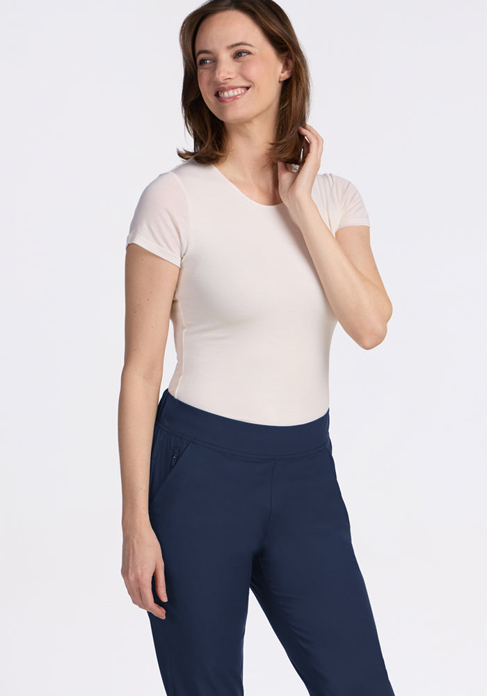 A woman with shoulder-length brown hair smiles while standing, wearing a short-sleeve white top and Woolx Nicole Ankle Pants in Deep Navy with a tapered-leg fit against a plain white background.
