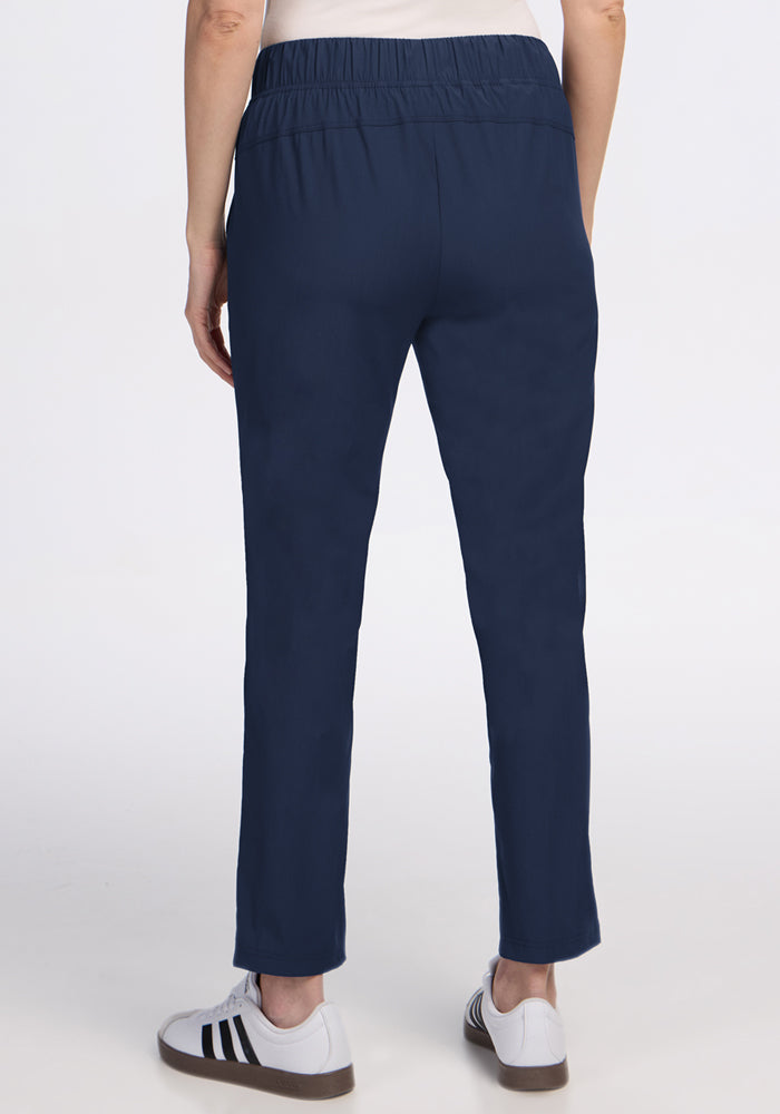 A person stands on a white background, back facing the camera, wearing Woolx Nicole Ankle Pants in Deep Navy with a tapered-leg fit and elastic waistband, paired with white sneakers featuring black stripes.