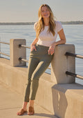 Smiling woman in a white tee and Woolx Nicole Ankle Pants - Sage Olive leans on a railing by the water in sunlight.