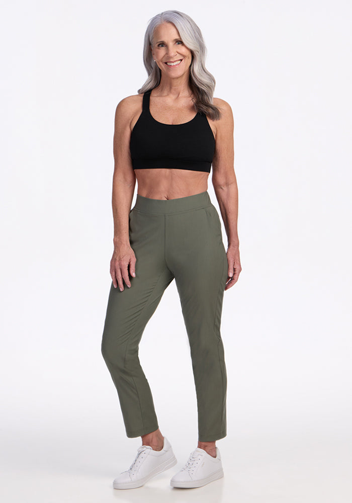 A smiling older woman with long gray hair wears the Woolx Nicole Ankle Pants in Sage Olive, a black sports bra, and white sneakers, standing confidently against a plain white background.