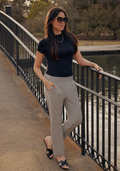 A woman in Woolx Nicole Ankle Pants - Vintage Khaki, sunglasses, and a black t-shirt stands on a bridge over a pond, one hand on the railing and the other in her pocket. Trees and water fill the background.