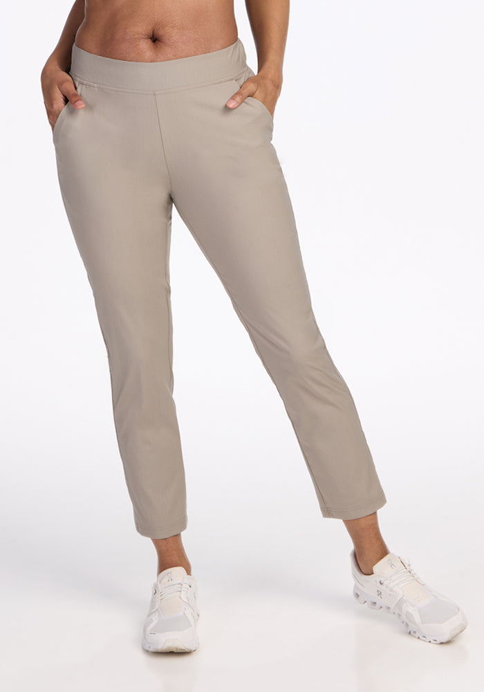 A person wears Woolx Nicole Ankle Pants in Vintage Khaki with white sneakers, standing hands-in-pockets against a plain white background. | Jade is 5’10”, wearing a size S
