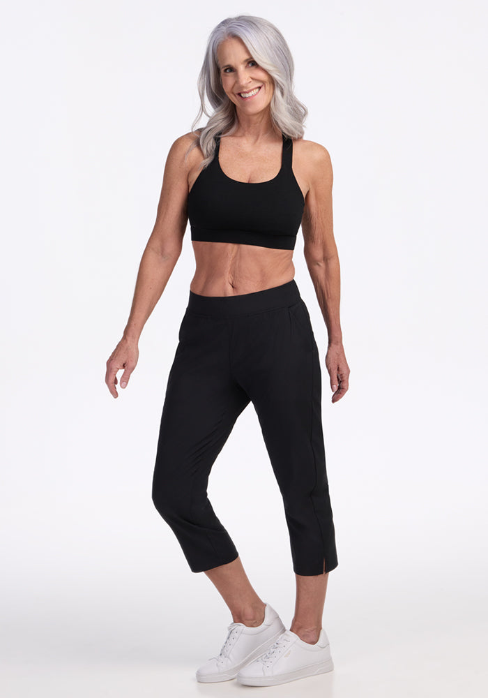 A smiling older woman with long gray hair confidently poses in a black sports bra, Woolx Nicole Crop Pants in black, and white sneakers against a plain white background.