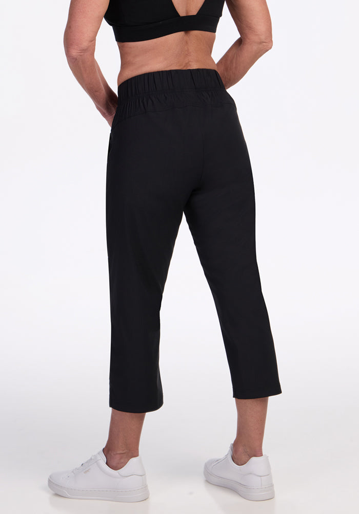 Someone stands facing away from the camera against a white background, dressed in Woolx Nicole Crop Pants - Black, a black sports bra, and white sneakers.