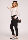 A woman smiles in a white long-sleeve top, Woolx Nicole Petite Ankle Pant in black, and white sneakers. She has sunglasses on her head, a black crossbody bag, and touches her hair with one hand. The background is plain white.