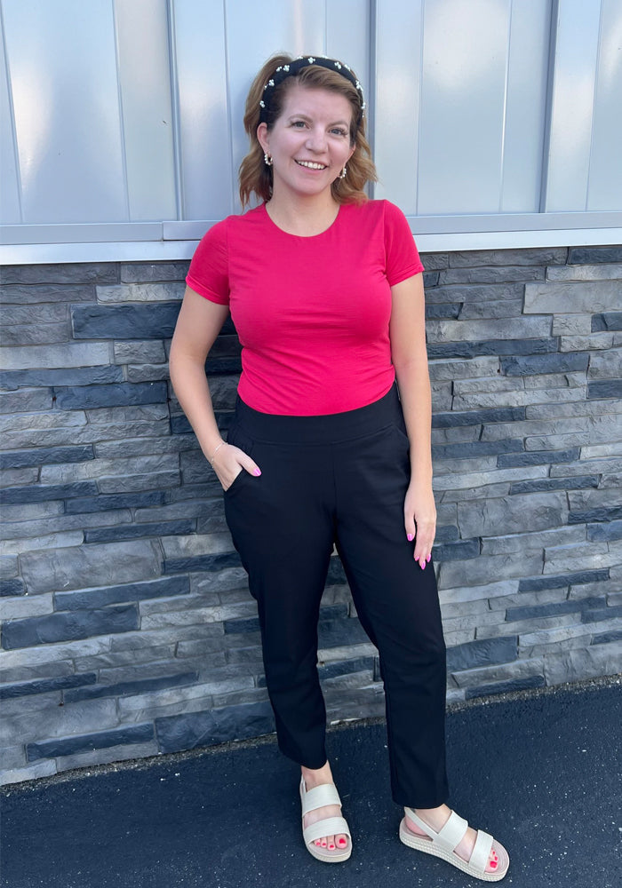 A woman smiles against a gray stone and metal wall, wearing a red t-shirt, Woolx Nicole Petite Ankle Pant in black, beige sandals, and a pearl headband. One hand is in her pocket as she stands on a blacktop surface.