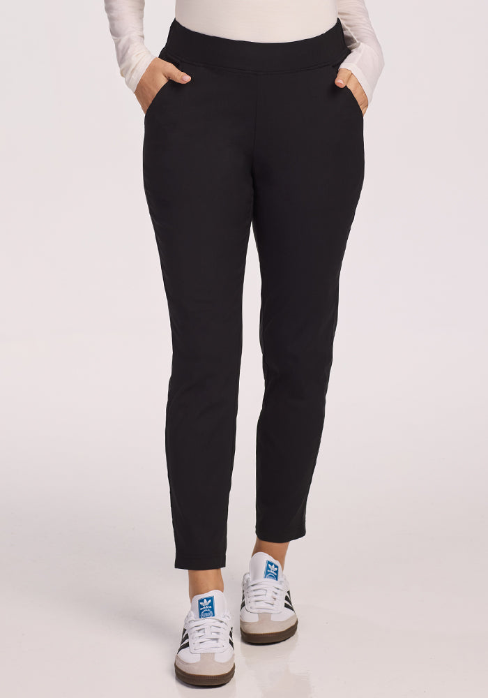 A person wearing Woolx Nicole Petite Ankle Pant in black stands with hands in pockets, paired with a long-sleeve white top and white sneakers with blue accents against a plain light background.