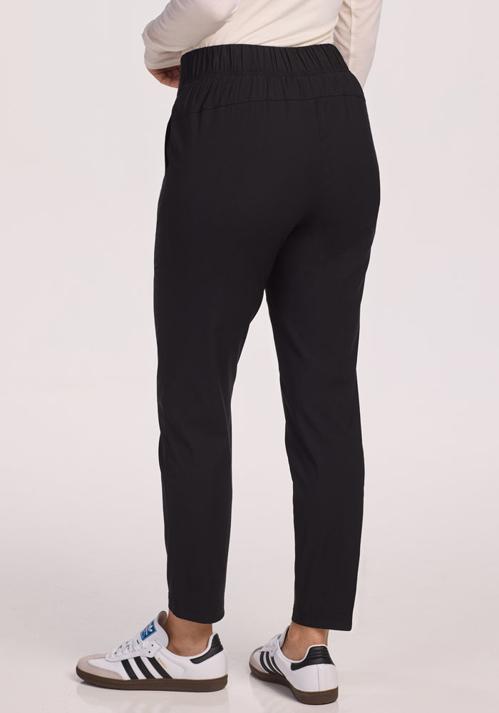 A person stands with their back to the camera against a light background, wearing Woolx Nicole Petite Ankle Pant in black and white sneakers with black stripes.