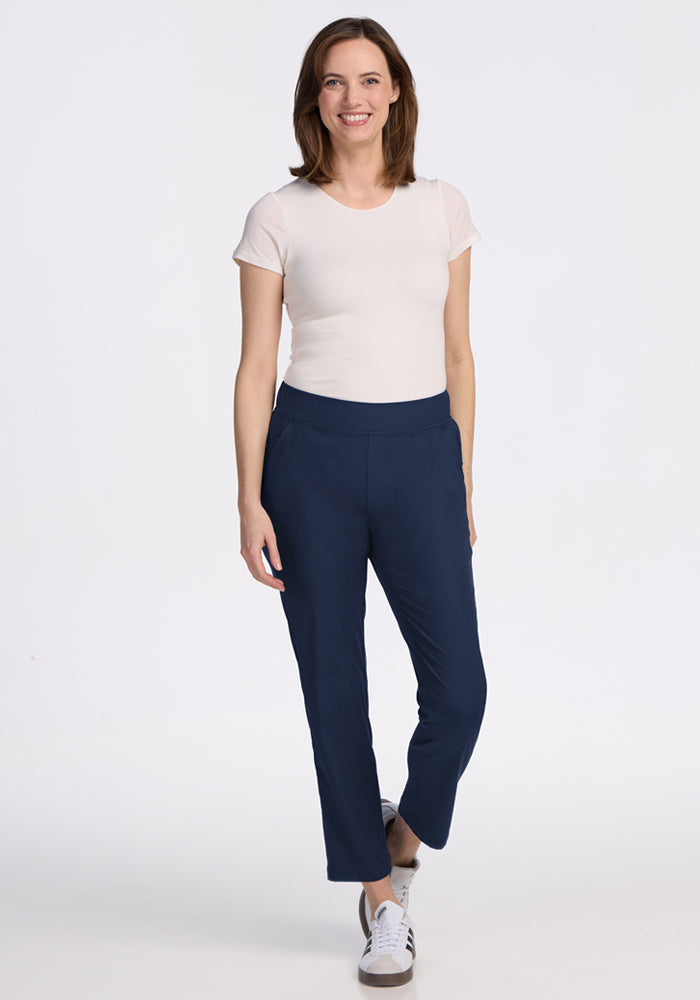 A woman with shoulder-length brown hair smiles against a white background, wearing a short-sleeved white shirt, Woolx Nicole Petite Ankle Pant in Deep Navy, and white sneakers.