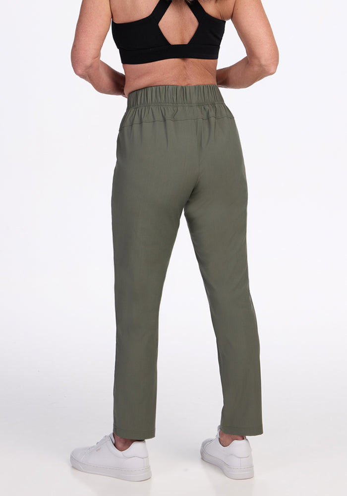 A person wearing Woolx Nicole Petite Ankle Pant in Sage Olive, a black sports bra, and white sneakers stands facing away from the camera against a plain white background.