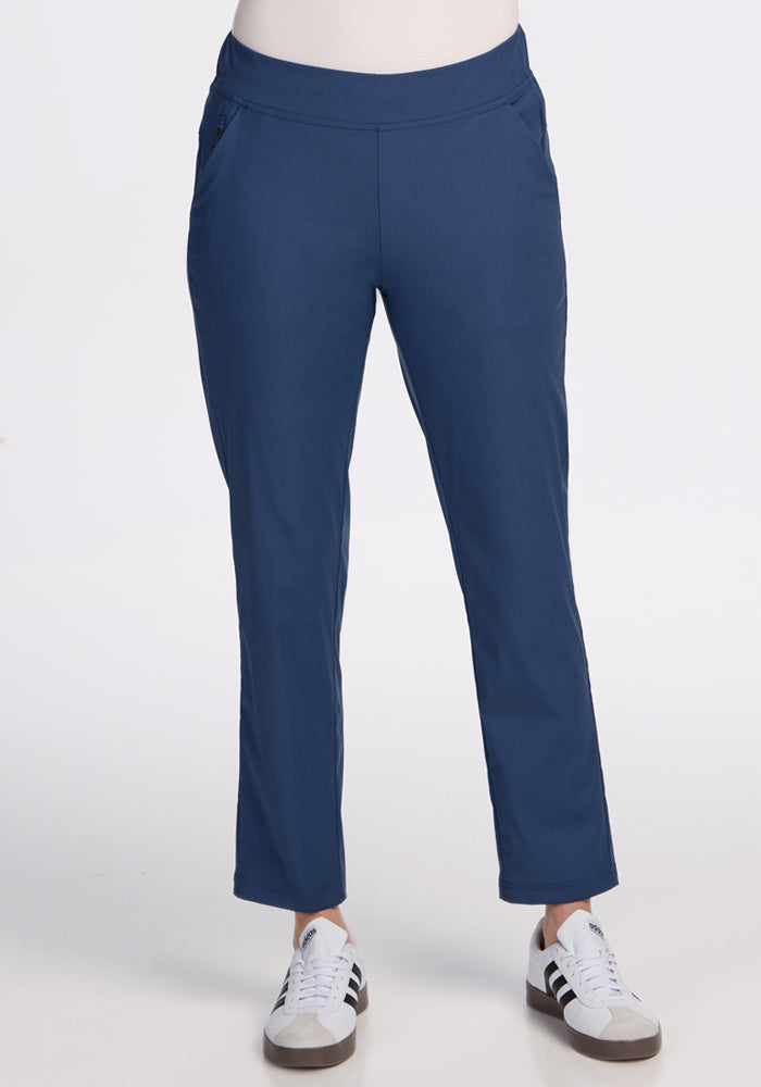 A person wears Woolx Nicole Petite Ankle Pant in Vintage Indigo and blue and white sneakers with black stripes, standing against a plain white background. Only the lower half of the body is shown.