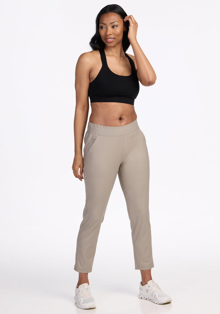 A woman poses against a white background in a black sports bra, white sneakers, and the Woolx Nicole Petite Ankle Pant in Vintage Khaki, with one hand near her head and a soft smile.