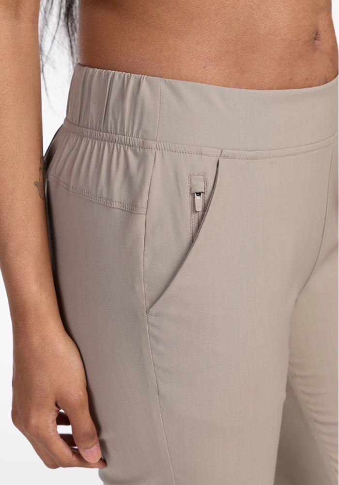 Close-up of a person wearing Woolx Nicole Petite Ankle Pant in Vintage Khaki, featuring a lightweight tapered design, elastic waistband, and zippered side pocket, with part of their bare midriff and left hand visible.